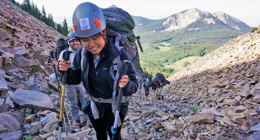 A student wearing a helmet and carrying a backpack uses trekking poles to navigate over a rocky area